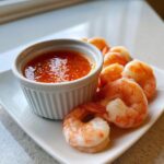 Plump, cooked shrimp arranged around a small white ramekin of bright orange shrimp cocktail sauce.