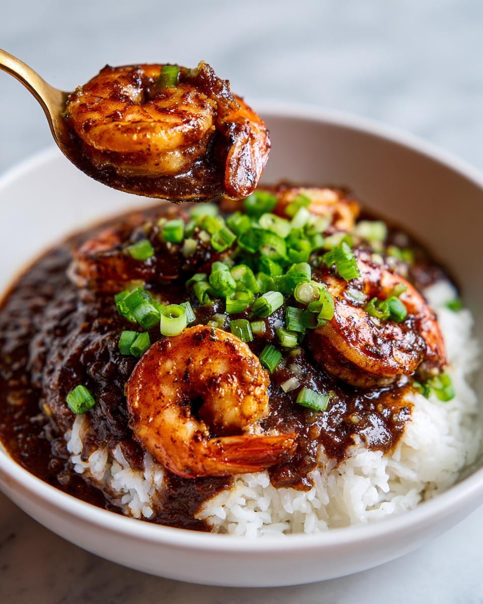 A spoonful of rich, dark shrimp étouffée featuring a seasoned shrimp is lifted above a bowl of white rice topped with green onions.