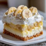 A close-up of a layered slice of banana cream pie recipe with a graham cracker crust, custard, whipped cream, and fresh banana slices.
