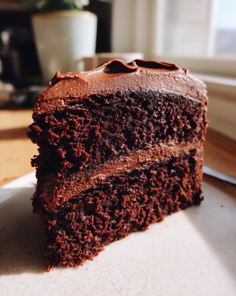 Close-up of a moist, dark slice from a chocolate cake recipe, featuring two layers and thick chocolate frosting.