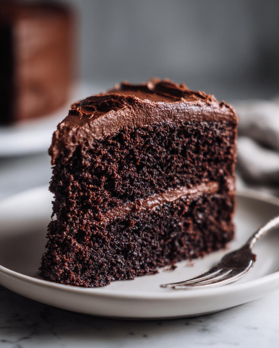 Close-up of a decadent slice of homemade chocolate cake with thick, dark chocolate frosting.