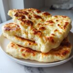 A stack of three fluffy, homemade naan bread pieces with golden-brown, slightly charred spots, served on a white plate.