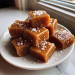 A close-up of soft, square caramels sprinkled with flaky sea salt, stacked on a small white plate.