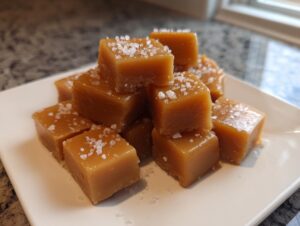 A close-up of soft, chewy caramels sprinkled with sea salt, stacked on a white serving plate.