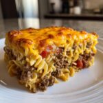 A hearty slice of sour cream beef noodle casserole topped with melted, browned cheddar cheese on a white plate.
