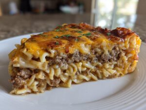 A thick slice of sour cream beef noodle casserole featuring layers of ground beef, pasta, and a bubbly, browned cheddar cheese topping.