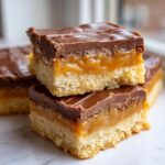 Two stacked homemade twix cookies bars showing a shortbread base, thick caramel layer, and chocolate topping.
