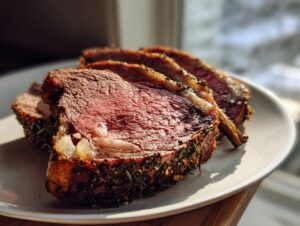 Close-up of several medium-rare slices of standing rib roast, crusted with herbs, resting on a white plate.