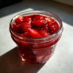 Close-up of bright red strawberry freezer jam with visible fruit chunks in a small glass jar.