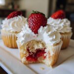 Close-up of a strawberry shortcake cupcake with a bite taken out, showing the strawberry filling and whipped cream topping.