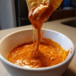 A tortilla chip is lifted from a bowl, showing long, gooey cheese strings stretching from the amazing chili cheese dip.