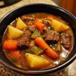 Close-up of a rich vegetable beef soup with chunks of beef, carrots, potatoes, and celery in a dark broth.