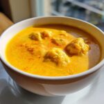 A close-up of a vibrant, orange-yellow cheese soup served in a white bowl, featuring several submerged dumplings.