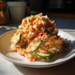 A close-up of a freshly prepared kani salad, featuring shredded imitation crab mixed with a creamy orange dressing and topped with thin slices of cucumber.