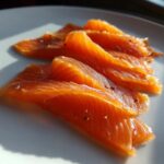 Close-up of vibrant orange slices of smoked salmon seasoned with pepper flakes on a white plate.