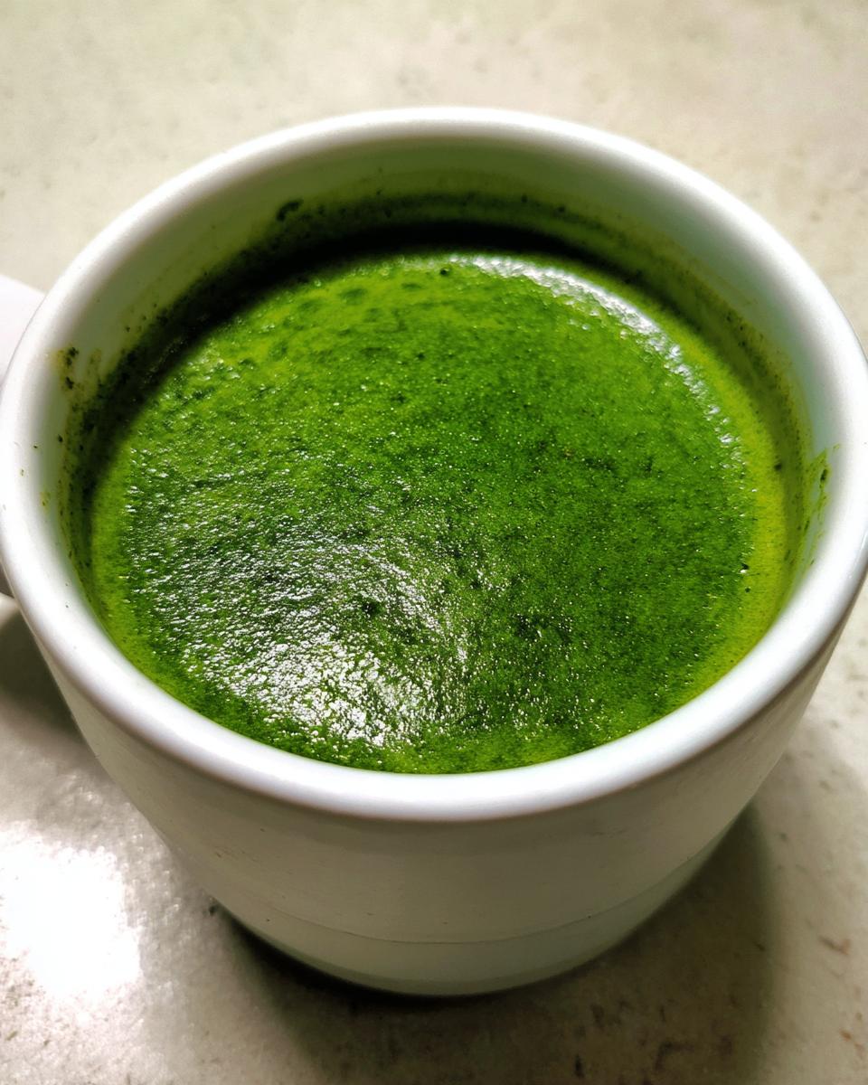 Close-up of vibrant green, frothy spinach soup served hot in a white mug.