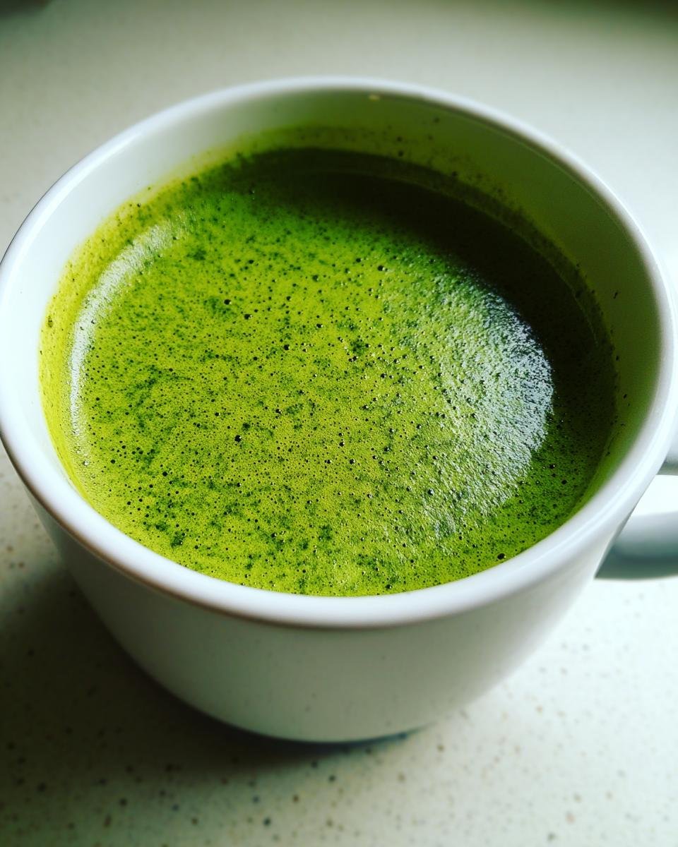 Close-up of vibrant green, frothy spinach soup served hot in a white ceramic mug.