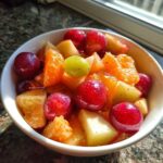 Close-up of a vibrant winter fruit salad featuring red grapes, orange segments, and apple chunks in a white bowl.