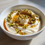 A close-up of creamy whipped ricotta dip swirled in a white bowl, drizzled with honey and topped with chopped pistachios.