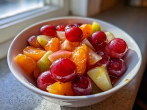 A close-up of a vibrant winter fruit salad featuring red grapes, orange segments, and light green apple chunks in a white bowl.
