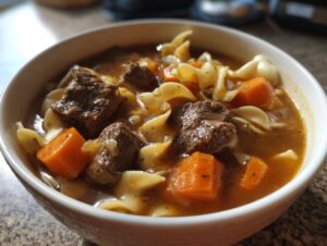 Close-up of a white bowl filled with rich beef noodle soup, featuring chunks of beef, carrots, and egg noodles.