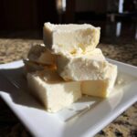A stack of creamy, pale yellow squares of homemade white chocolate fudge resting on a white plate.
