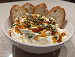 A white bowl filled with creamy ricotta dip, drizzled with honey and topped with fresh basil, served with toasted baguette slices.