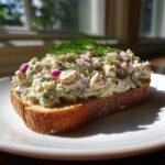 A generous serving of creamy salmon salad piled high on a slice of toasted bread, garnished with fresh dill.