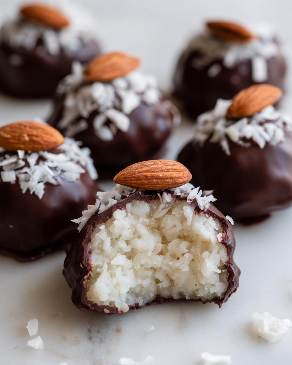 A close-up shot of several chocolate-covered almond joy truffles, one of which is bitten open revealing the white coconut filling.