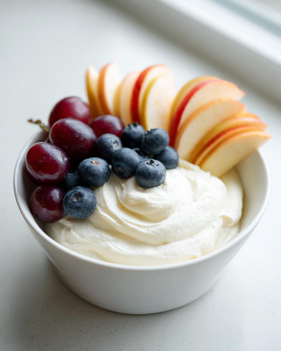 A white bowl filled with creamy fruit dip topped with fresh blueberries, red grapes, and sliced apples.