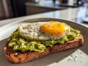 A close-up of delicious avocado toast topped with a sunny-side-up egg, pepper, and sea salt.