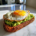 A close-up of delicious avocado toast on dark bread, topped with mashed avocado and a sunny-side-up egg seasoned with pepper.