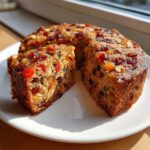 A thick slice cut from a rich, dense fruit cake recipe, loaded with colorful dried fruits and nuts, sitting on a white plate.