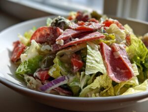 A close-up view of a vibrant grinder salad featuring lettuce, salami, red onion, olives, and creamy dressing.