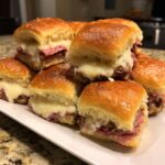 A close-up stack of warm, cheesy grinder sliders on a white platter.