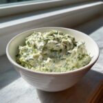 A close-up of creamy, green pickle dip mixed with fresh dill served in a white bowl.
