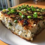 A square slice of baked rice dish, known as sushi bake, showing a golden-brown, crispy top garnished with fresh green onions.