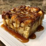 A close-up of a moist slice of apple bread pudding generously topped with dripping caramel sauce.
