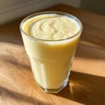 A tall glass filled with a thick, creamy yellow banana smoothie, sitting on a sunlit wooden table.