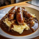 Close-up of three browned sausages served over creamy mashed potatoes, smothered in rich onion gravy, classic bangers and mash.