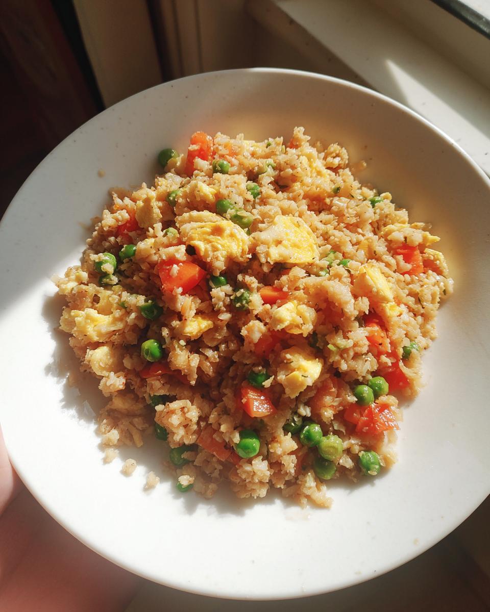 A white bowl filled with homemade cauliflower fried rice mixed with scrambled egg, bright green peas, and diced carrots.