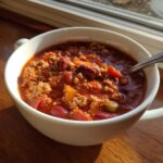 A close-up of a white mug filled with rich, hearty high protein chili, featuring ground meat, kidney beans, and vegetables.