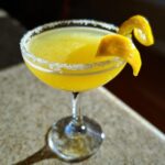 A bright yellow lemon drop cocktail served in a coupe glass with a sugared rim and a lemon twist garnish.