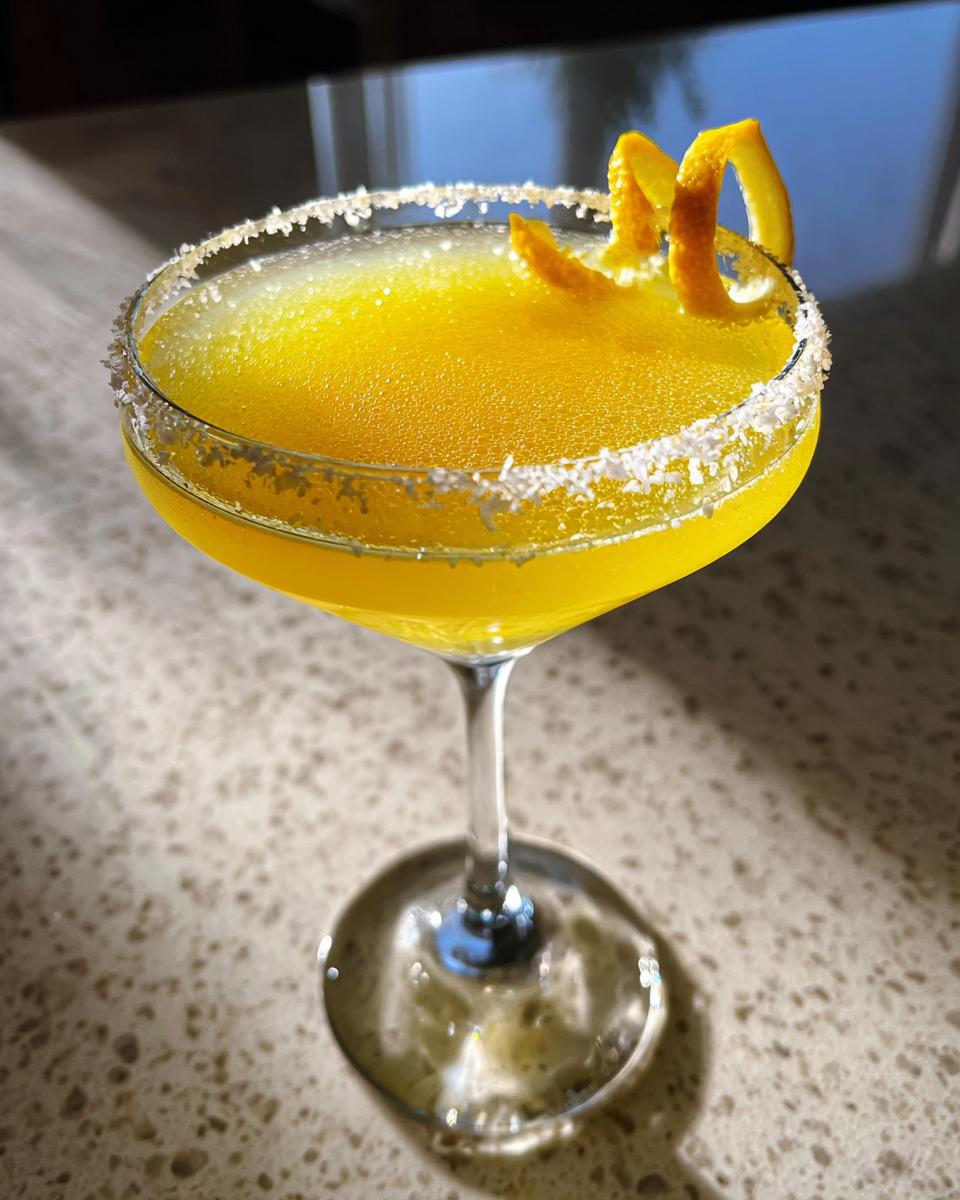 A bright yellow lemon drop cocktail in a coupe glass with a sugared rim and orange peel garnish.