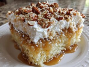 A close-up of a slice of butter pecan praline poke cake with whipped topping and dripping caramel sauce.