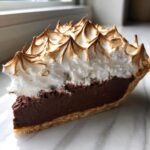 A close-up slice of chocolate meringue pie showing a rich dark filling and tall, beautifully toasted meringue peaks.