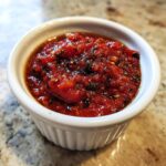 Close-up of a small white ramekin filled with chunky, vibrant red pizza sauce seasoned with dark herbs.