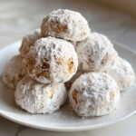A close-up of a stack of amazing cinnamon sugar snowball cookies heavily dusted with powdered sugar on a white plate.