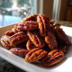A close-up shot of a pile of shiny, dark brown candied pecans served on a white dish.