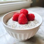 A white bowl filled with creamy coconut chia pudding topped with fresh, bright red raspberries.